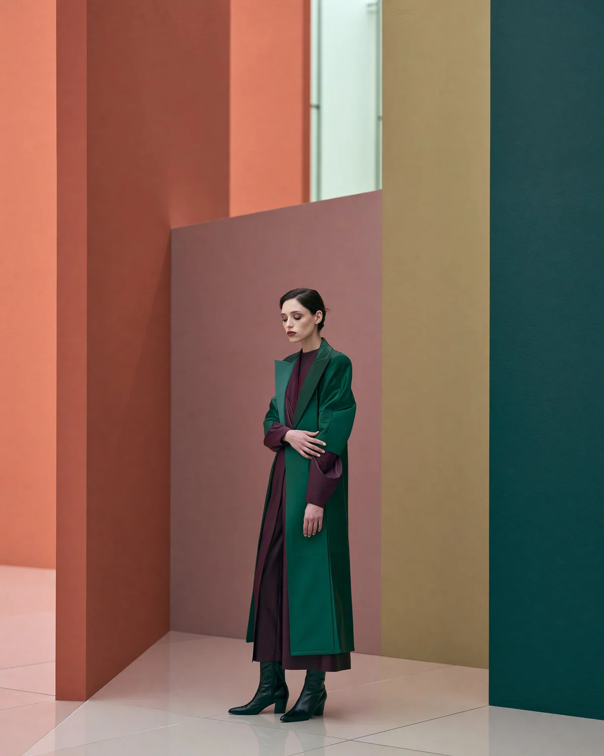 Full-body fashion editorial in a green coat against warm color-blocked walls.