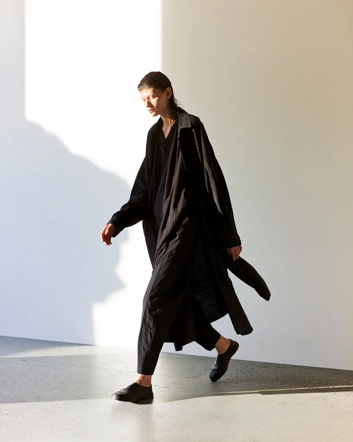 Full-body black fashion silhouette walking through a graphic sunlit shadow.