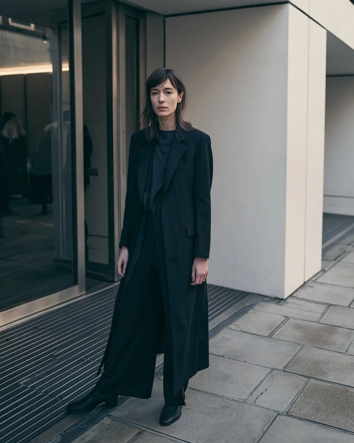 Full-body street-style fashion portrait in black tailoring near a glass entry.