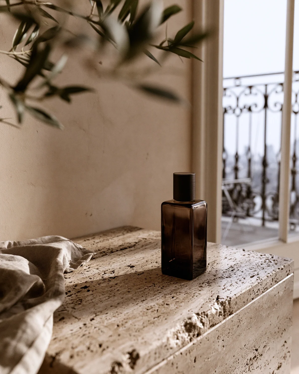 Dark unbranded fragrance bottle on travertine in soft balcony light.