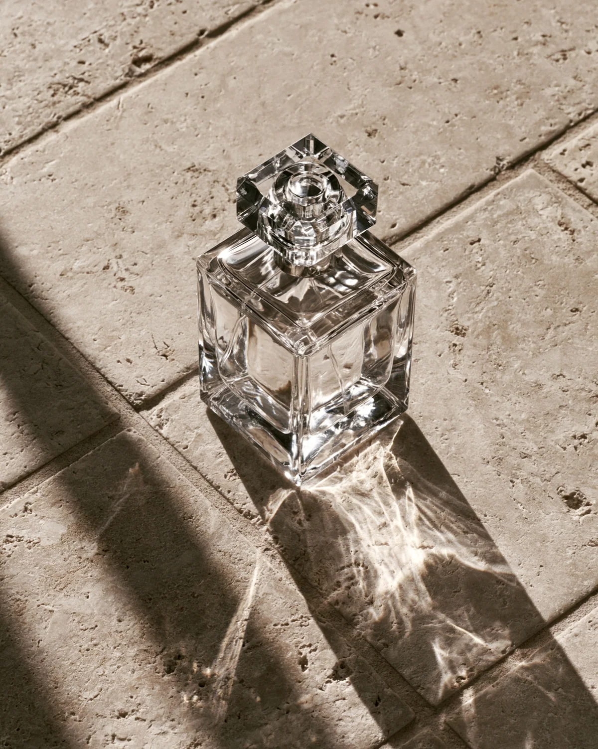 Clear perfume bottle on sunlit limestone with refracted shadows.