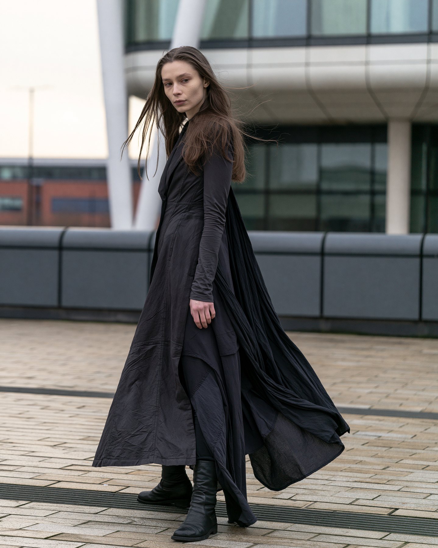 Full-body fashion editorial model in a flowing black coat on an urban terrace.