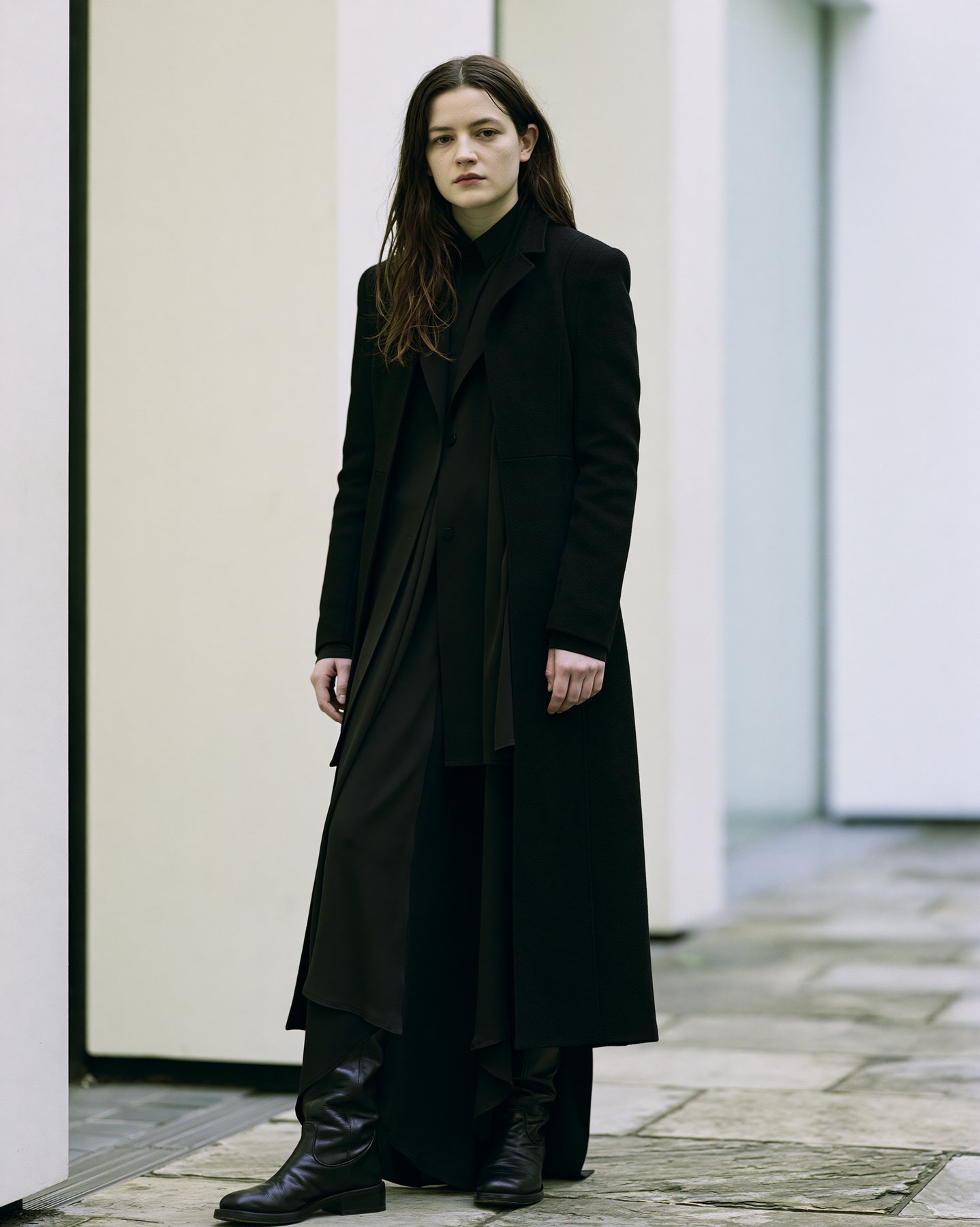 Full-body fashion editorial model in a long black coat against a pale architectural backdrop.