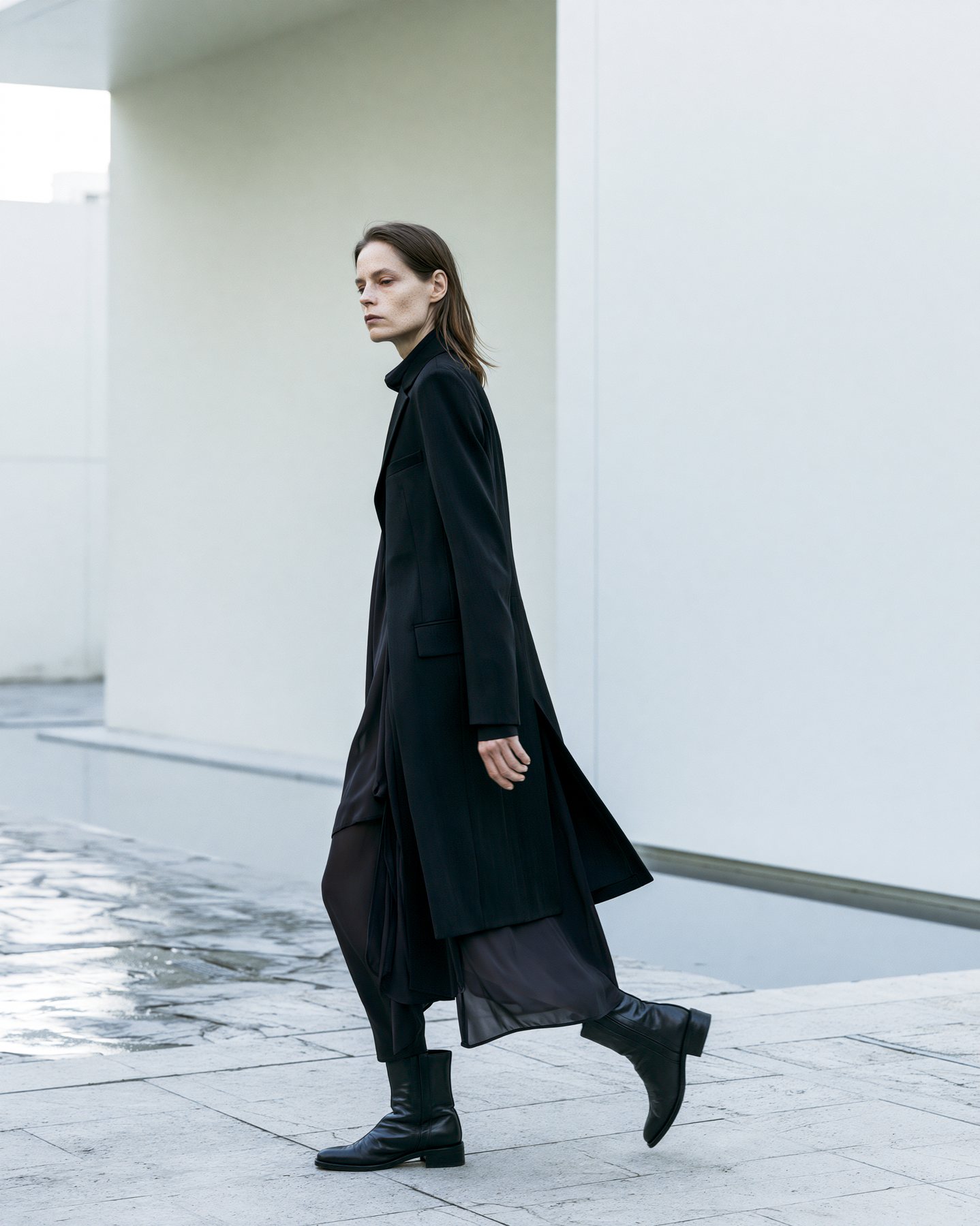 Full-body fashion model walking past pale modern architecture in a black coat.