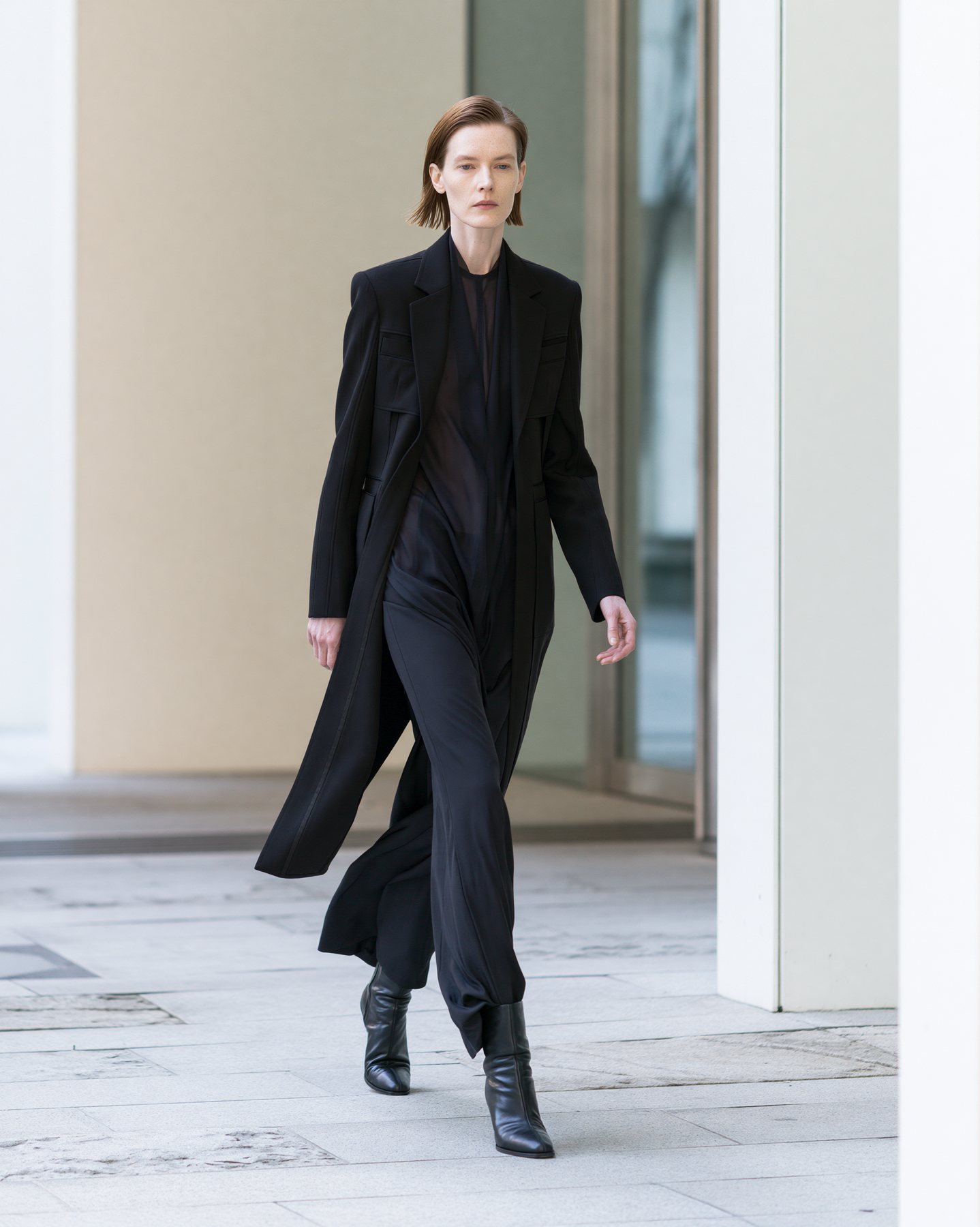 Full-body black tailored fashion look photographed in a clean architectural corridor.