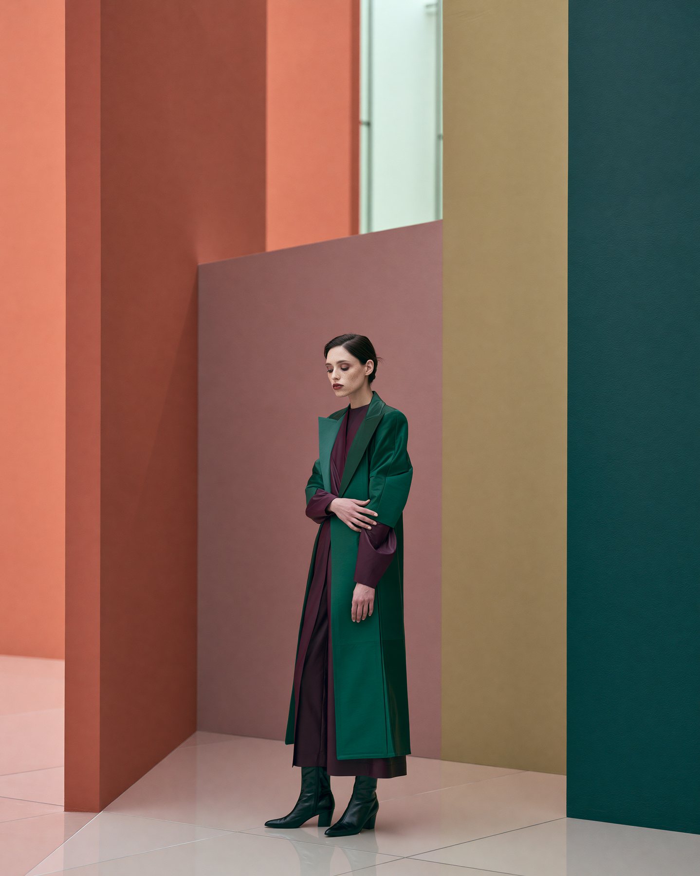 Full-body fashion editorial in a green coat against warm color-blocked walls.