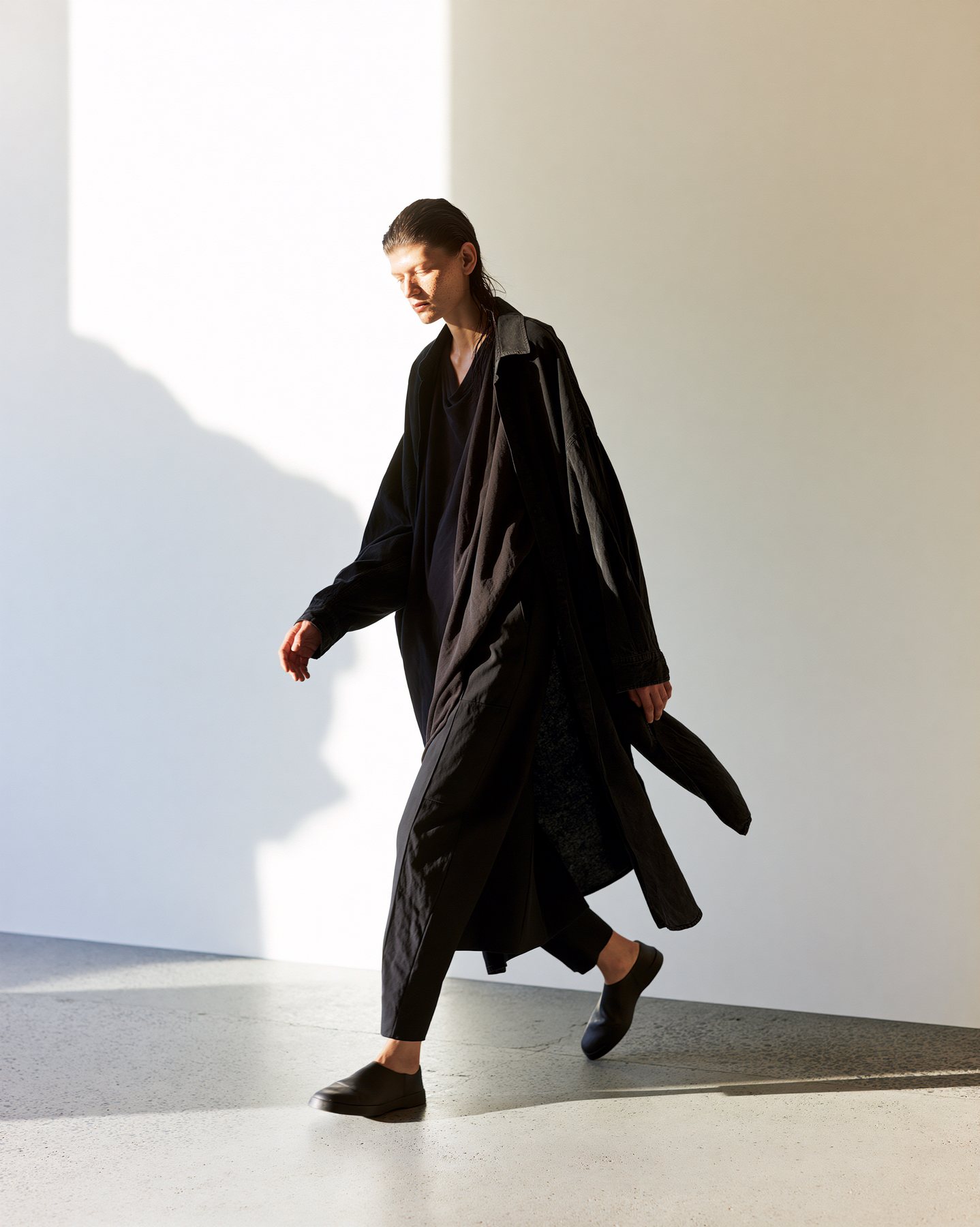 Full-body black fashion silhouette walking through a graphic sunlit shadow.