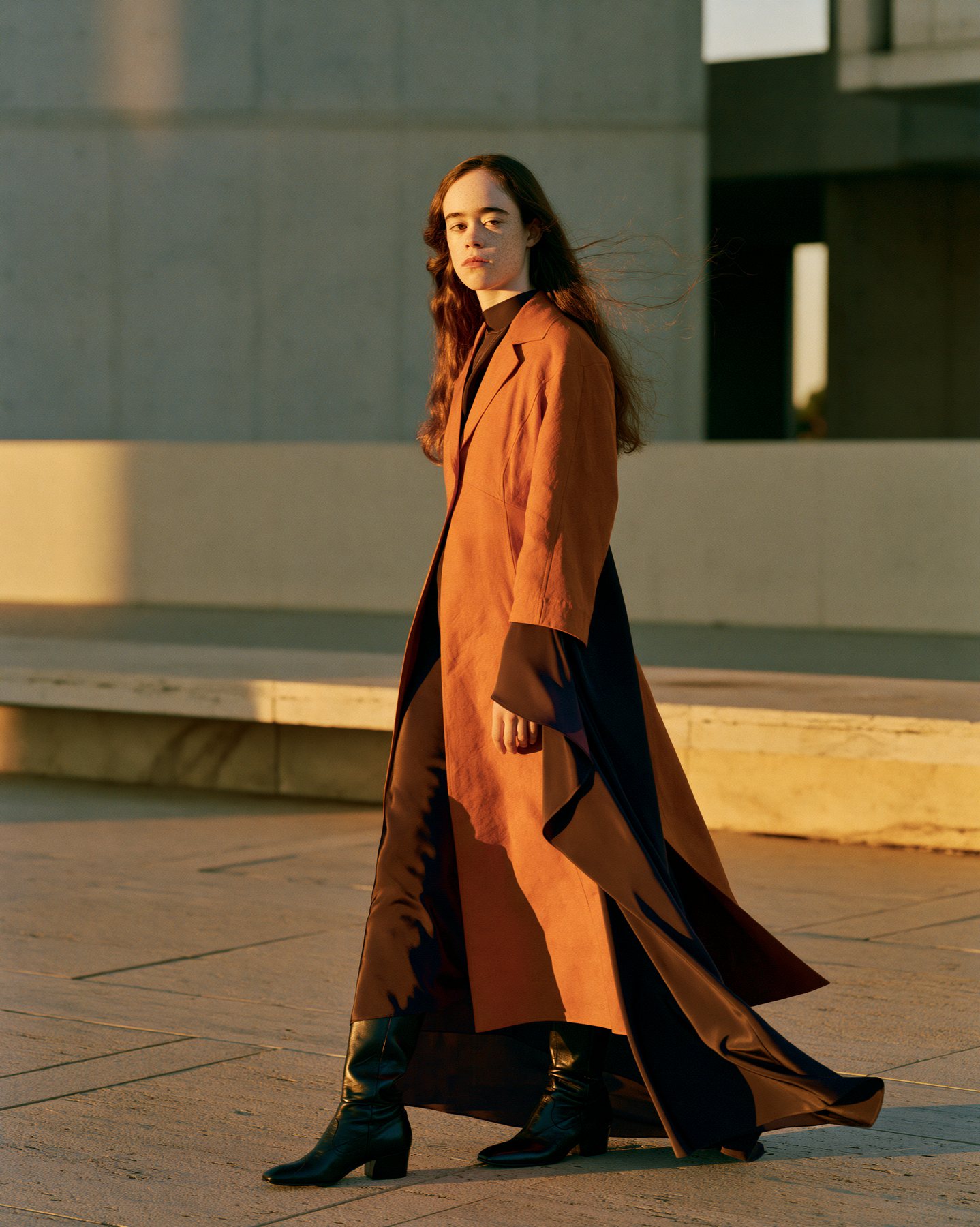 Full-body fashion editorial in a long amber coat during golden urban light.