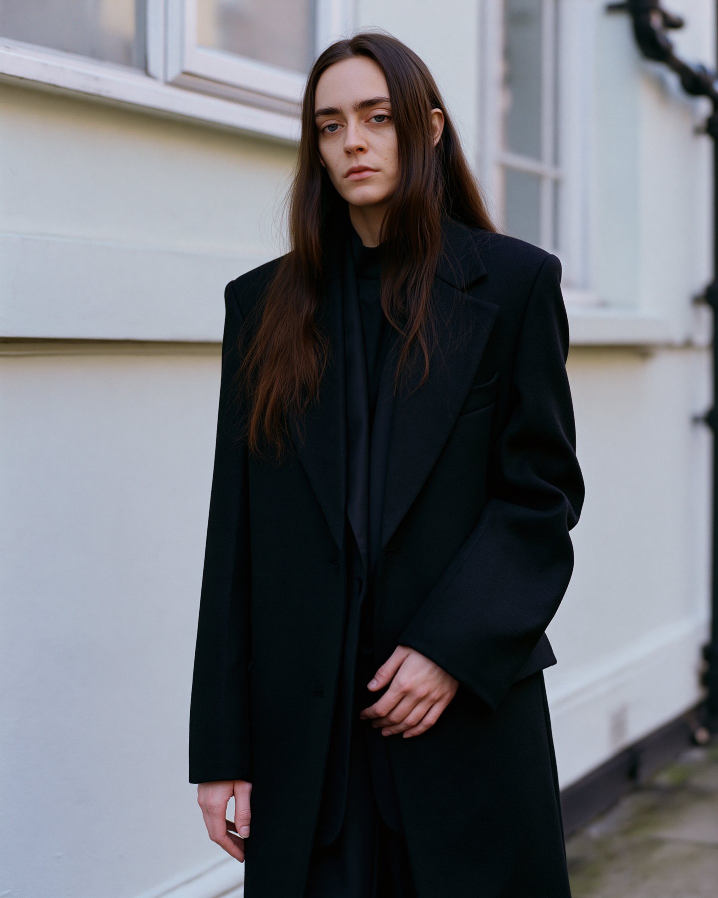Realistic fashion portrait in black tailoring against a pale urban facade.