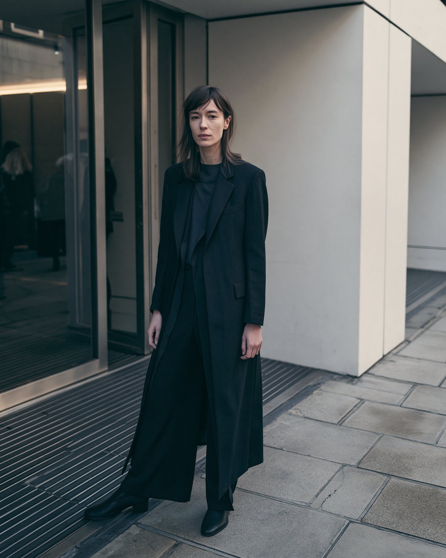 Full-body street-style fashion portrait in black tailoring near a glass entry.