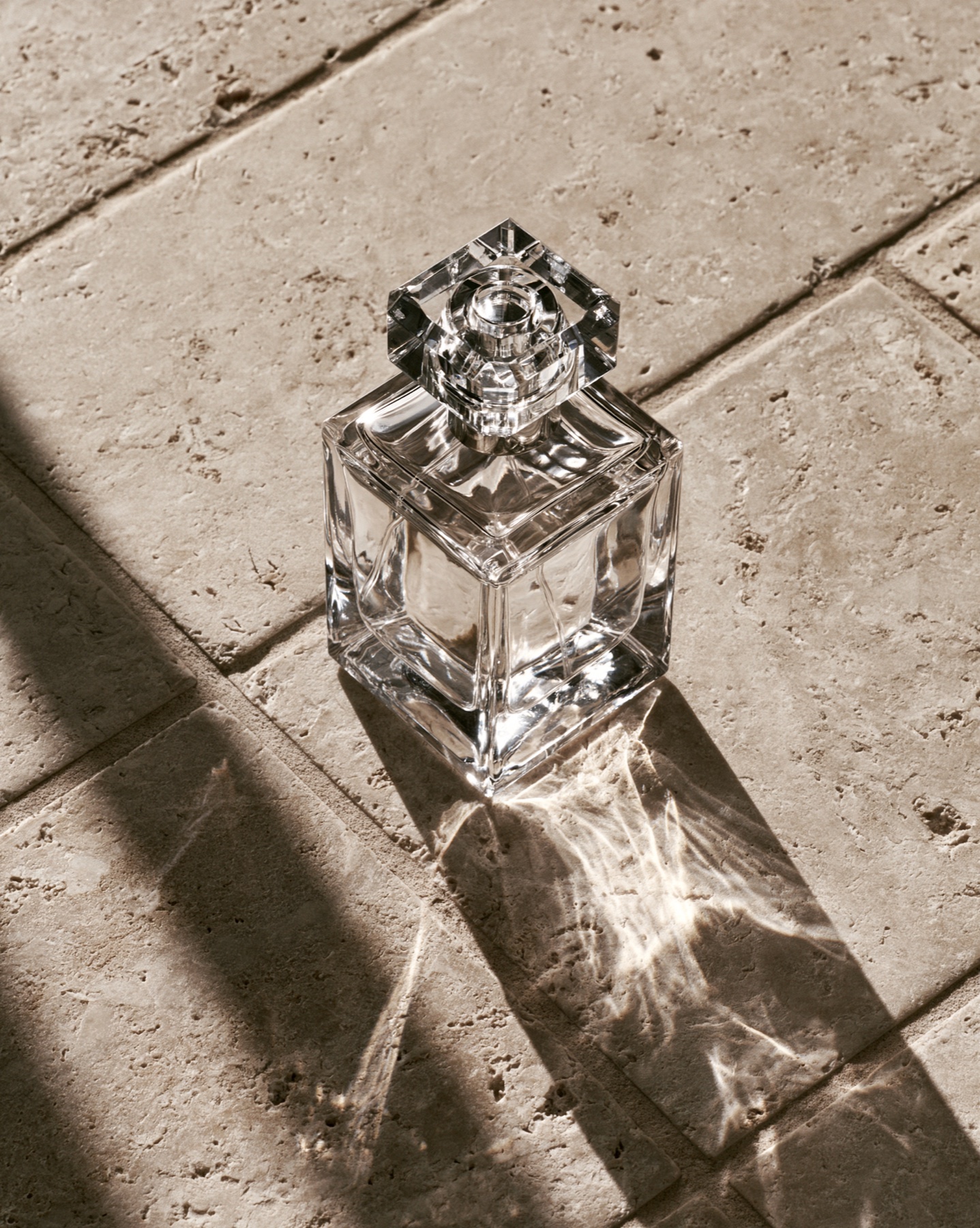 Clear perfume bottle on sunlit limestone with refracted shadows.